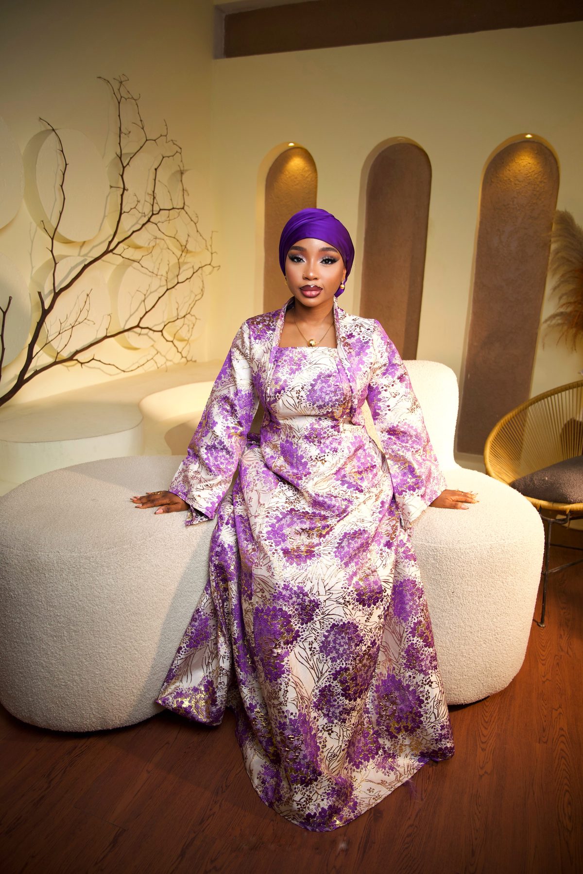 Bridal client in a purple silk gele and purple-and-gold floral gown, seated in a warm arched studio setting